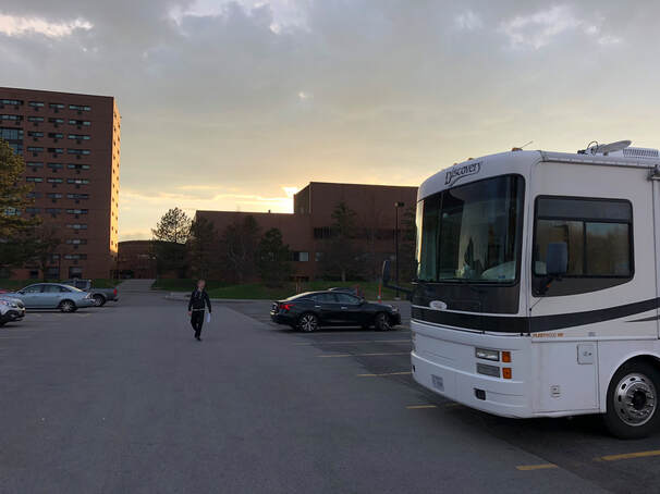White motorhome on right side parked on NTID parking lot with several tall buildings against sunset, and a blond haired white teenager walking towards the camera.
